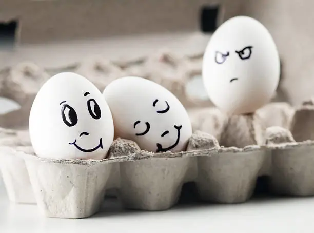 Sur cette image ludique, trois œufs dans une boîte à œufs semblent représenter une scène de relations humaines. Deux œufs sont joyeusement collés l'un à l'autre, s'étreignant avec tendresse, tandis que le troisième, relégué à l'arrière, arbore une expression mécontente, presque envieuse, de voir les deux autres si heureux ensemble. C'est une illustration amusante des dynamiques relationnelles et des émotions complexes que peuvent susciter les schémas d'imperfection.