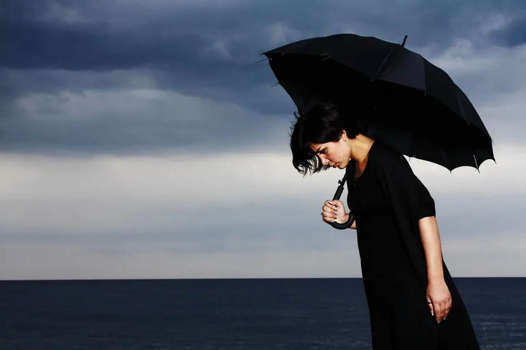 Une femme vêtue de noir se tient seule, un parapluie noir comme bouclier contre la pluie des émotions qui la submerge. Sa tête baissée trahit le poids immense qu'elle porte sur ses épaules. Dans le ciel, des nuages gris reflètent la tempête intérieure qui fait rage en elle, amplifiant le sentiment d'isolement et d'abandon.